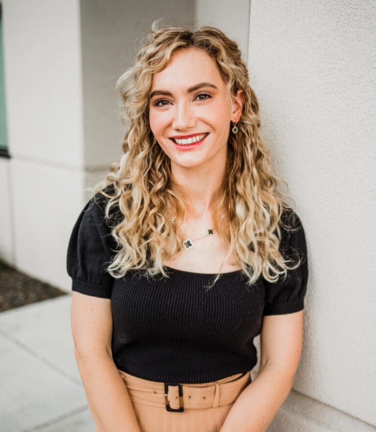 Woman with curly blonde hair wearing a black top and beige skirt, smiling while leaning against a wall.