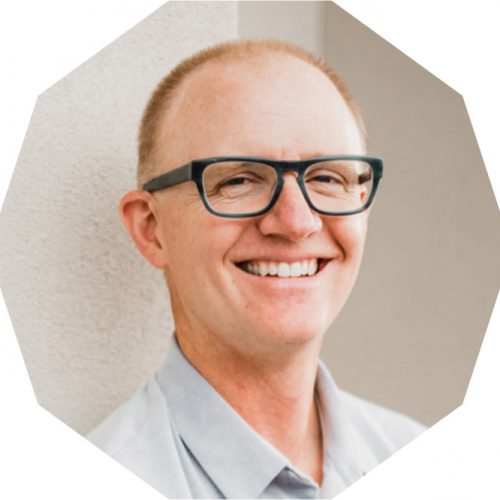 A man with glasses and a light shirt is smiling while standing against a light-colored wall. The image is cropped into an octagonal shape.