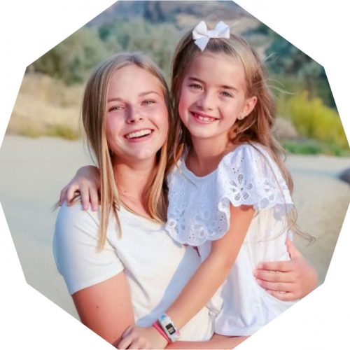 Two smiling girls pose together outdoors. The older girl holds the younger, who wears a white dress with a bow in her hair. They both appear happy, set against a blurred natural background.